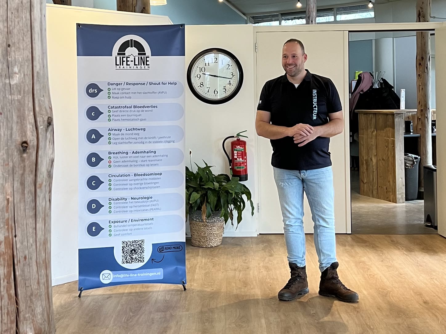 Een glimlachende instructeur van Life-Line Trainingen staat naast een banner met EHBO-protocollen
